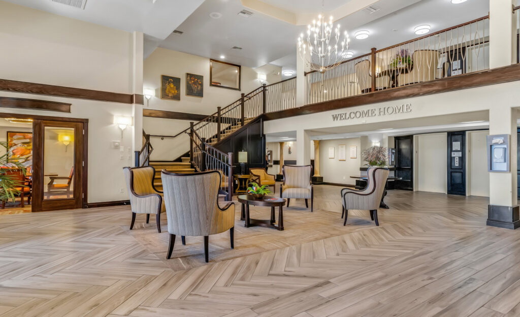 A lobby room with six beige sofa chairs arranged in a circle around a coffee table, and a staircase leading to the second floor, where the golden letters say "Welcome Home."
