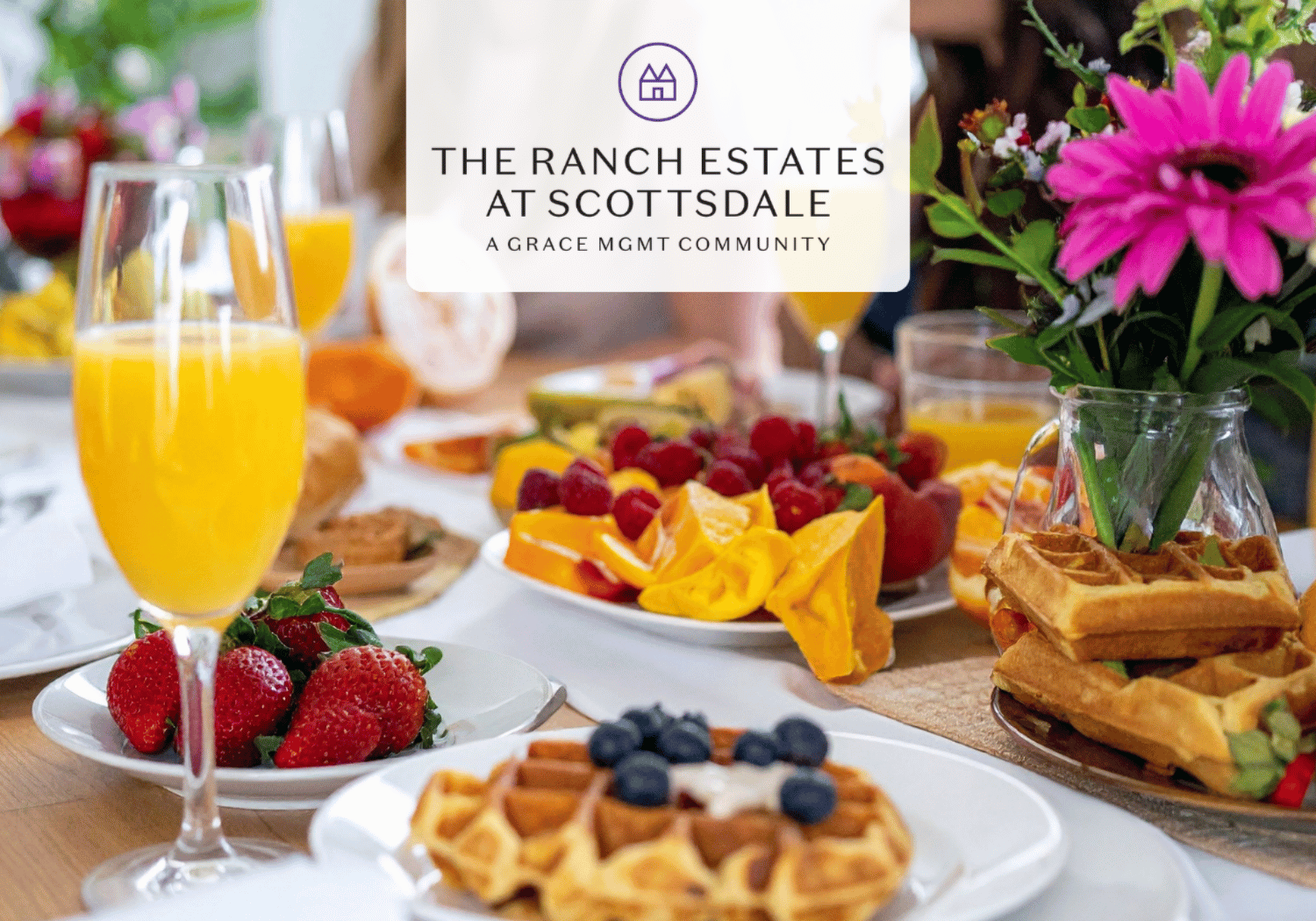 Table set for brunch with waffles, berries, orange juice, and a vase of pink flowers; a white banner in the center reads 'The Ranch Estates at Scottsdale, a Grace Mgmt Community'